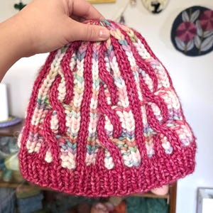 May include: A hand-knitted beanie in a vibrant fuchsia color with a cable knit design. The hat features vertical stripes and interwoven patterns in white, pink, and pastel hues. This handmade hat is a cozy accessory for cold weather.