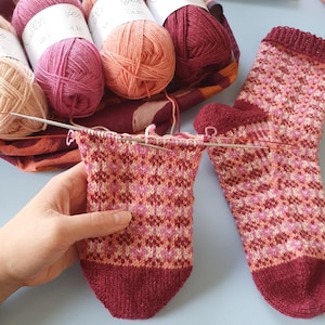 May include: A partially knitted sock in shades of pink, peach, and burgundy, with a geometric pattern. Knitting needles are inserted into the work, and several balls of yarn in coordinating colors are visible in the background. The sock is on a light blue surface.