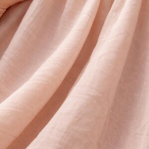 May include: Close-up of soft, light pink fabric with visible texture and folds. The fabric appears to be lightweight and flowing, possibly linen or a similar material. A hand gently touches the fabric, highlighting its delicate nature.
