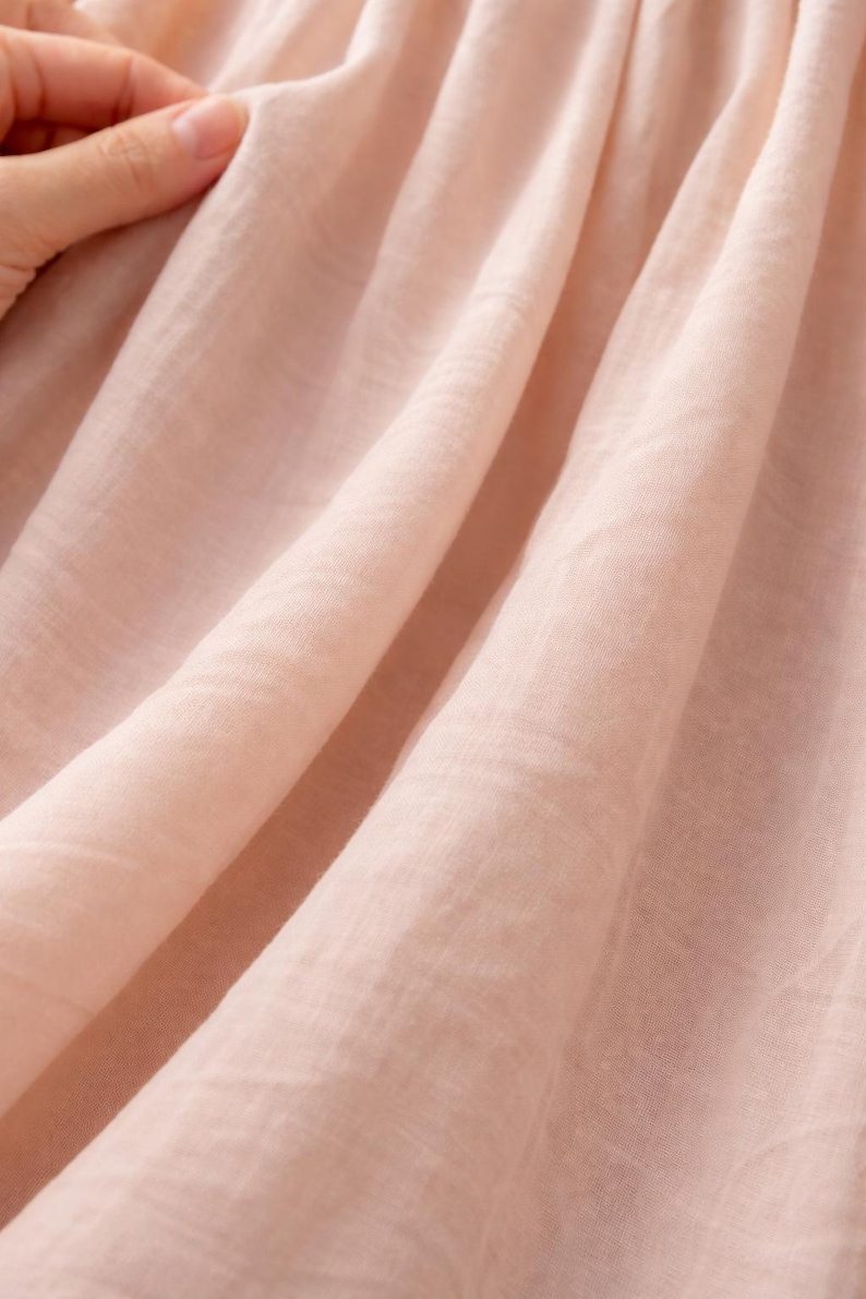 May include: Close-up of soft, light pink fabric with visible texture and folds. The fabric appears to be lightweight and flowing, possibly linen or a similar material. A hand gently touches the fabric, highlighting its delicate nature.