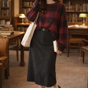 May include: A woman in a library setting, wearing a red and black plaid sweater, a black skirt, and black shoes. She carries a white tote bag and holds a book. The background features bookshelves and reading lamps.