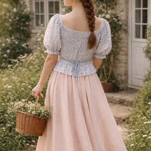 May include: A woman in a floral print top and a long pink skirt carries a woven basket filled with white flowers. The outfit has a light blue tie at the waist. The woman has long red hair in a braid.
