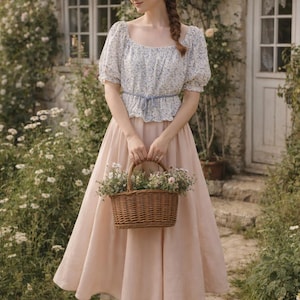 May include: A woman in a light blue floral top and a long, blush-colored skirt holds a woven basket filled with flowers. She wears brown flats. The scene is set in front of a stone cottage with a thatched roof, surrounded by greenery.