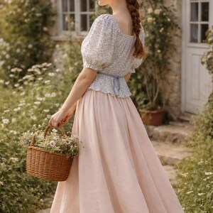 May include: A woman in a light pink skirt and a floral print top carries a woven basket filled with flowers. The outfit is paired with brown shoes. The background features a cottage with a thatched roof and lush greenery, creating a romantic, pastoral scene.