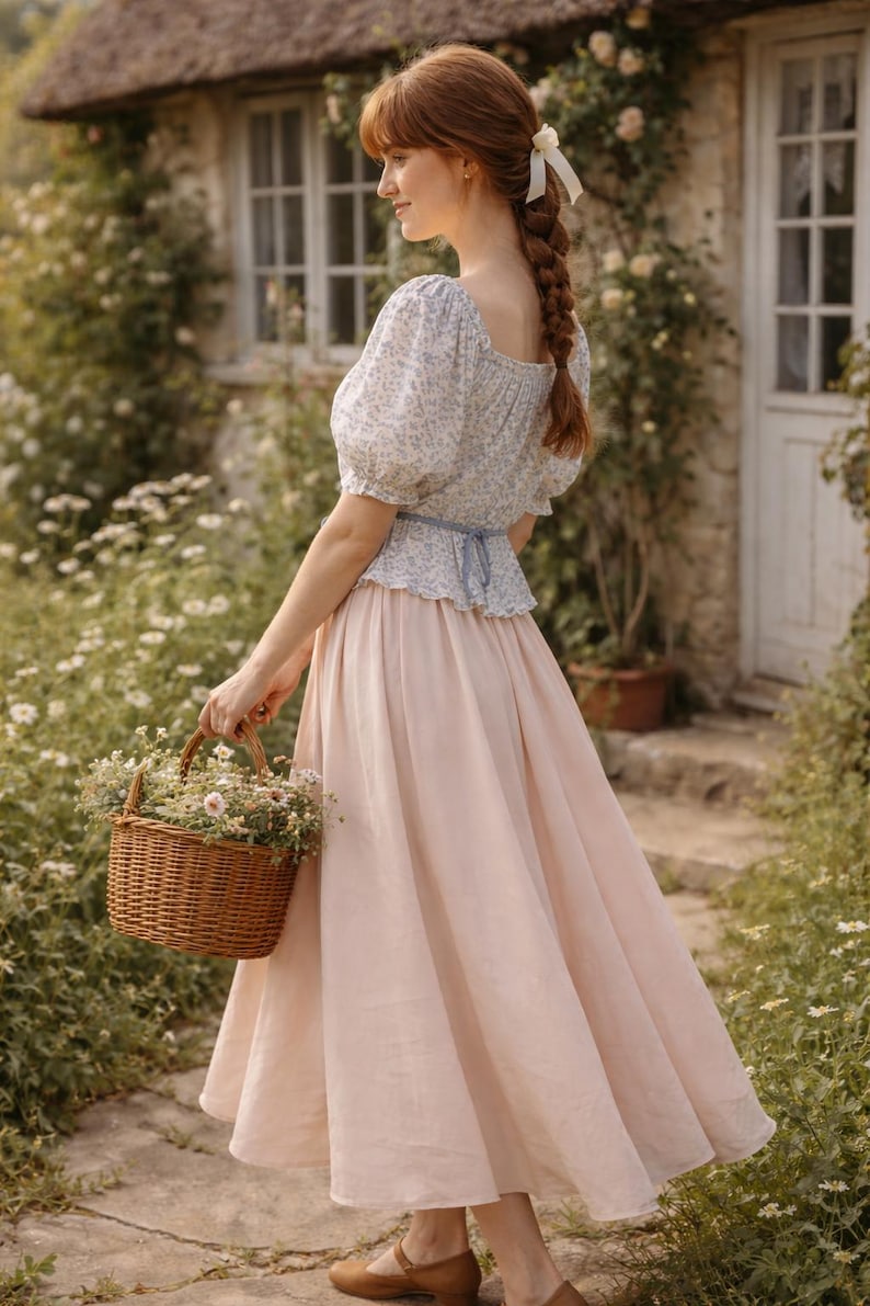 May include: A woman in a light pink skirt and a floral print top carries a woven basket filled with flowers. The outfit is paired with brown shoes. The background features a cottage with a thatched roof and lush greenery, creating a romantic, pastoral scene.
