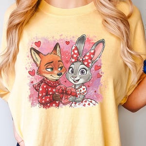 May include: Yellow t-shirt with a cartoon image of a fox and a rabbit, both holding a heart-shaped gift. The fox is in a red sweater with white hearts, and the rabbit wears a dress with heart details. The background is pink with heart accents.
