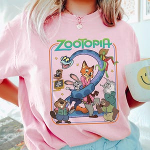 Puede incluir: Camiseta rosa claro con un dise&ntilde;o colorido de Zootopia, con personajes y el n&uacute;mero 2. El dise&ntilde;o es de estilo retro. Una taza blanca con una carita sonriente amarilla se sostiene en la mano.