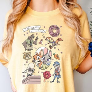 Puede incluir: Camiseta amarilla con personajes de dibujos animados de Zootopia. El dise&ntilde;o incluye a Nick Wilde, Judy Hopps y otros personajes, junto con el texto "You can be whatever you wish to be." La camiseta tambi&eacute;n tiene el texto "THE BIG DONUT."