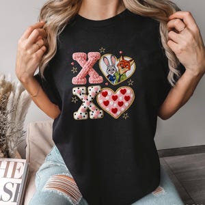 May include: Black t-shirt featuring embroidered designs. The design includes the letters "XO" in pink knit and black and white polka dot patterns, along with heart-shaped images of cartoon characters. The word "BLACK" is printed at the bottom.