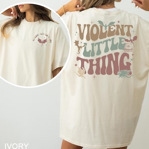 May include: Ivory-colored t-shirt with the text "VIOLENT LITTLE THING" in a retro font. The design includes dragon illustrations in various colors. The shirt has a crew neck and short sleeves.