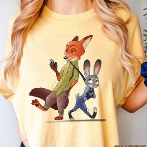 Może przedstawiać: Ż&oacute;łta koszulka z kresk&oacute;wkową ilustracją Nicka Wilde'a, lisa, i Judy Hopps, kr&oacute;lika, z Zootopii. Nick trzyma telefon, a Judy idzie na smyczy. Słowo "BUTTER" jest wydrukowane w prawym dolnym rogu.