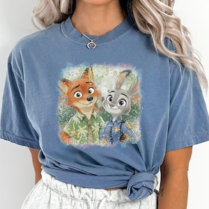 以下が含まれることがあります： ズートピアのニック・ワイルドとジュディ・ホップスのカラフルなグラフィックが特徴のブルージーンズカラーのTシャツ。画像には、下部に「BLUE JEAN」というテキストが含まれています。シャツは半袖で、クルーネックです。