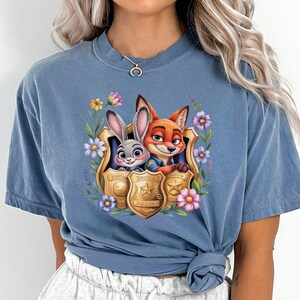 Pu&ograve; includere: T-shirt blu jeans con una grafica colorata di Judy Hopps e Nick Wilde di Zootopia, incorniciata da fiori e distintivi della polizia. Il testo "Zootopia Police" &egrave; visibile sui distintivi. La maglietta ha una vestibilit&agrave; rilassata.