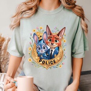 May include: Light green t-shirt featuring a cartoon image of a blue bunny and a red fox in police uniforms inside a gold badge with the word "POLICE". The badge is surrounded by flowers. The shirt is a light green color.
