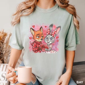 May include: Pale green t-shirt with a cartoon fox and rabbit, both holding a heart-shaped gift box, with hearts around them. The fox is in a red sweater, and the rabbit wears a dress with heart patterns. The shirt has the word "BAY" printed on the bottom right.