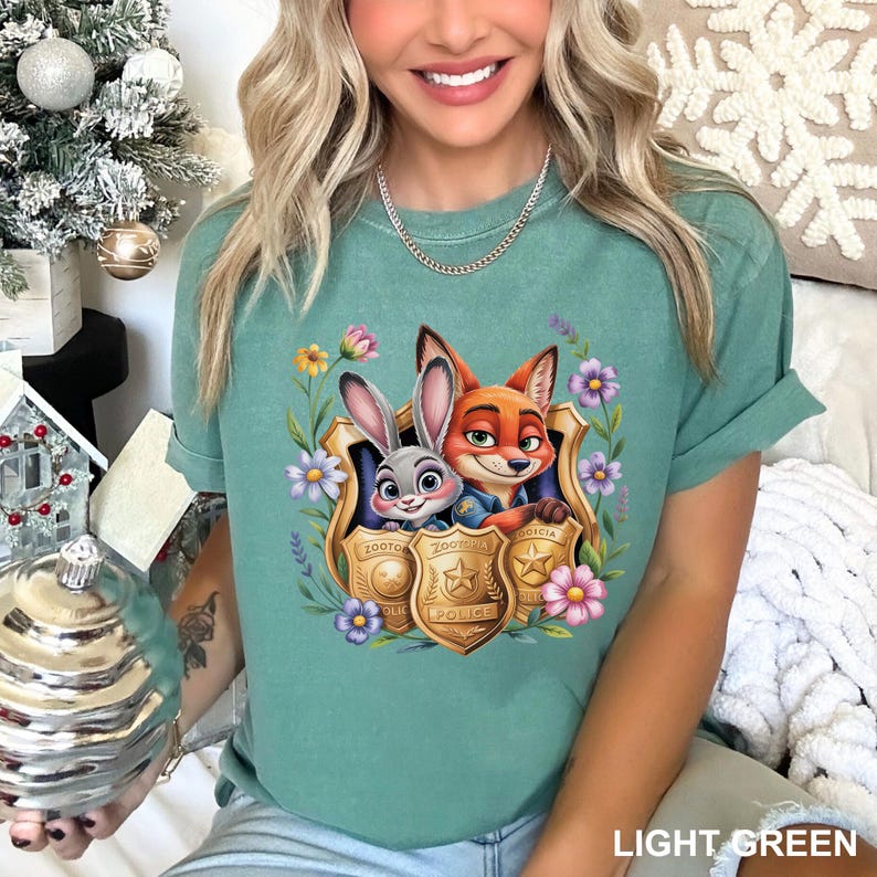 Pu&ograve; includere: T-shirt verde chiaro con una grafica colorata di Judy Hopps e Nick Wilde di Zootopia, incorniciata da fiori e distintivi della polizia. Il testo "Zootopia Police" &egrave; visibile sui distintivi. La maglietta &egrave; di colore verde chiaro.