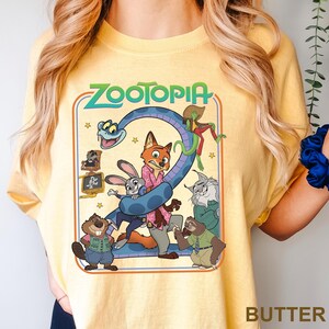Puede incluir: Camiseta de color mantequilla con un gr&aacute;fico colorido de la pel&iacute;cula animada Zootopia. El dise&ntilde;o incluye el t&iacute;tulo de la pel&iacute;cula, una serpiente azul y varios personajes de animales. La camiseta es de uso casual y c&oacute;modo.