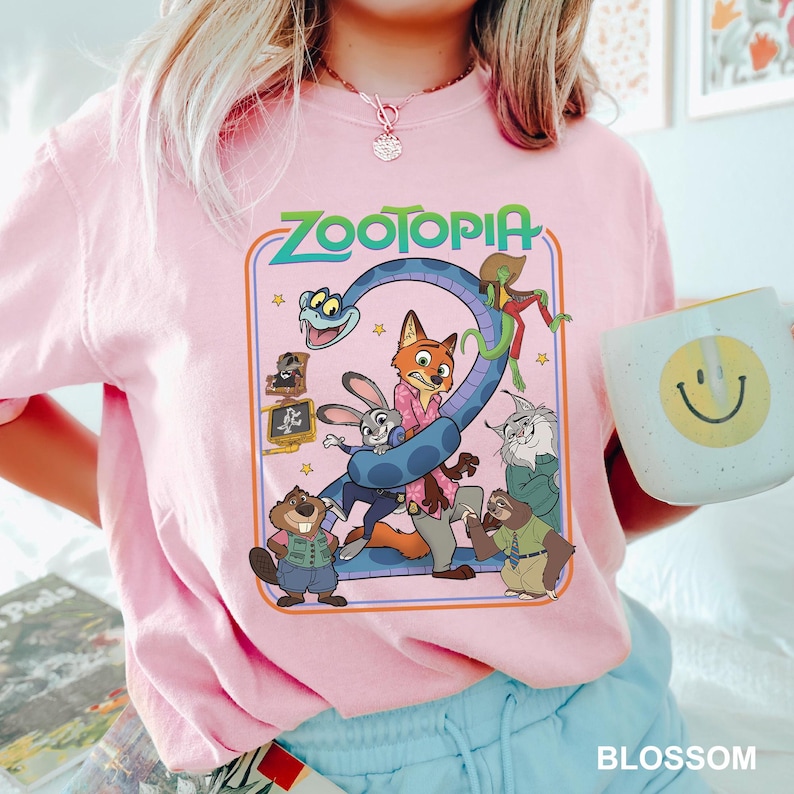 Puede incluir: Camiseta rosa claro con un dise&ntilde;o colorido de Zootopia, con personajes y el n&uacute;mero 2. El dise&ntilde;o es de estilo retro. Una taza blanca con una carita sonriente amarilla se sostiene en la mano.