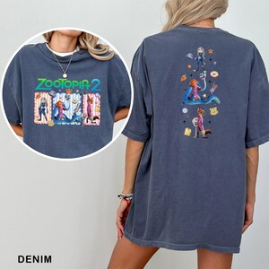 May include: Denim blue t-shirt featuring the text "ZOOTOPIA 2" with cartoon characters. The back of the shirt has a vertical design with more cartoon characters and food items. The shirt is a relaxed fit.