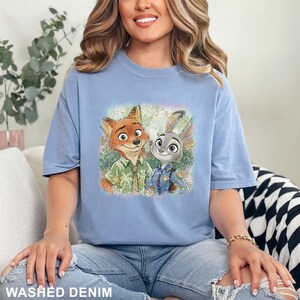 以下が含まれることがあります： 水色のTシャツ。映画「ズートピア」のキャラクター、ニック・ワイルドとジュディ・ホップスのカラフルなグラフィックがプリントされています。Tシャツの下部には「WASHED DENIM」の文字がプリントされています。