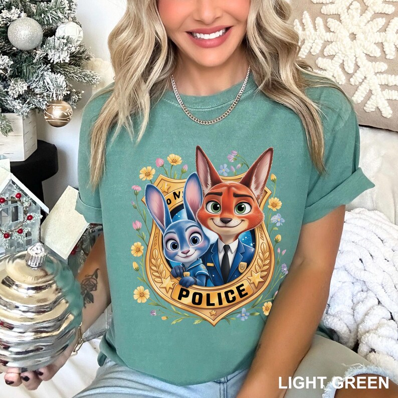 May include: Light green t-shirt featuring a cartoon image of Judy Hopps and Nick Wilde from Zootopia inside a gold police badge. The badge is surrounded by yellow flowers and green leaves. The word "POLICE" is printed on the badge.