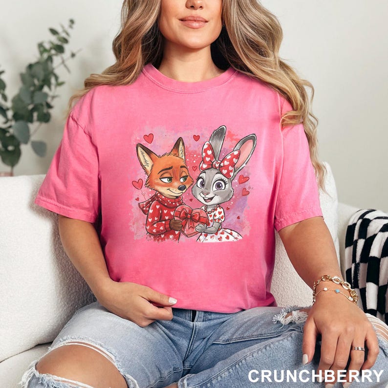 May include: A pink t-shirt with a cartoon fox and rabbit, each holding a heart-shaped gift. The fox is in a red scarf, the rabbit has a red bow. Small red hearts are scattered around. The word "CRUNCHBERRY" is printed at the bottom right of the image.