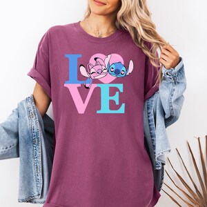 May include: A maroon t-shirt with the word "LOVE" in blue and pink block letters. The "O" is replaced by a pink heart with cartoon characters. A denim jacket is draped over the shoulders.