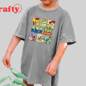 Pu&ograve; includere: Una maglietta grigia con una grafica colorata di Toy Story che presenta Woody, Buzz Lightyear e altri personaggi. Il testo "Toy Story" &egrave; stampato in un riquadro giallo.