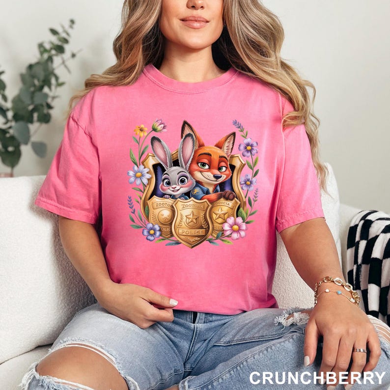 Pu&ograve; includere: Una maglietta rosa con un'illustrazione di Judy Hopps e Nick Wilde di Zootopia, incorniciati da fiori e un distintivo della polizia. La maglietta &egrave; di colore rosa unito.