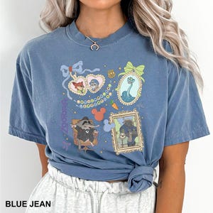 Puede incluir: Camiseta azul vaquero con un gr&aacute;fico colorido de personajes e iconos enmarcados. El dise&ntilde;o incluye im&aacute;genes de un zorro, un conejo, una serpiente y un mapache. La palabra "zootopia" est&aacute; impresa verticalmente en el lado izquierdo de la camiseta.
