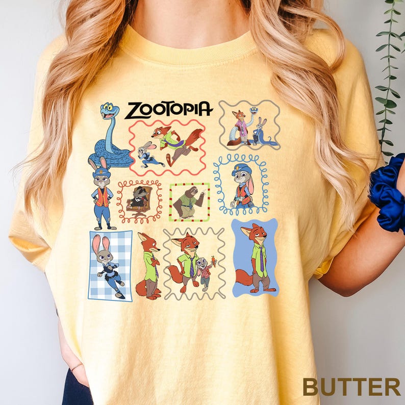 May include: Butter-colored t-shirt featuring a colorful graphic of characters from the animated film "Zootopia." The design includes various characters in framed illustrations, with the film's title at the top. The word "BUTTER" is printed at the bottom.