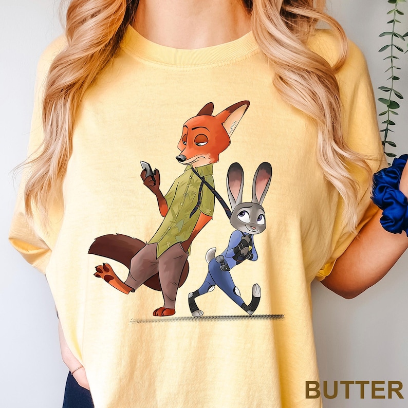 Może przedstawiać: Ż&oacute;łta koszulka z kresk&oacute;wkową ilustracją Nicka Wilde'a, lisa, i Judy Hopps, kr&oacute;lika, z Zootopii. Nick trzyma telefon, a Judy idzie na smyczy. Słowo "BUTTER" jest wydrukowane w prawym dolnym rogu.