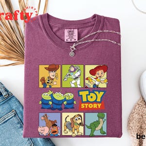 Pu&ograve; includere: Maglietta viola con una grafica di personaggi del film Toy Story. I personaggi includono Woody, Buzz Lightyear, Jessie, i tre piccoli alieni verdi, Rex e Mr. Potato Head. Il testo "Toy Story" &egrave; stampato in lettere bianche su uno sfondo rosso.