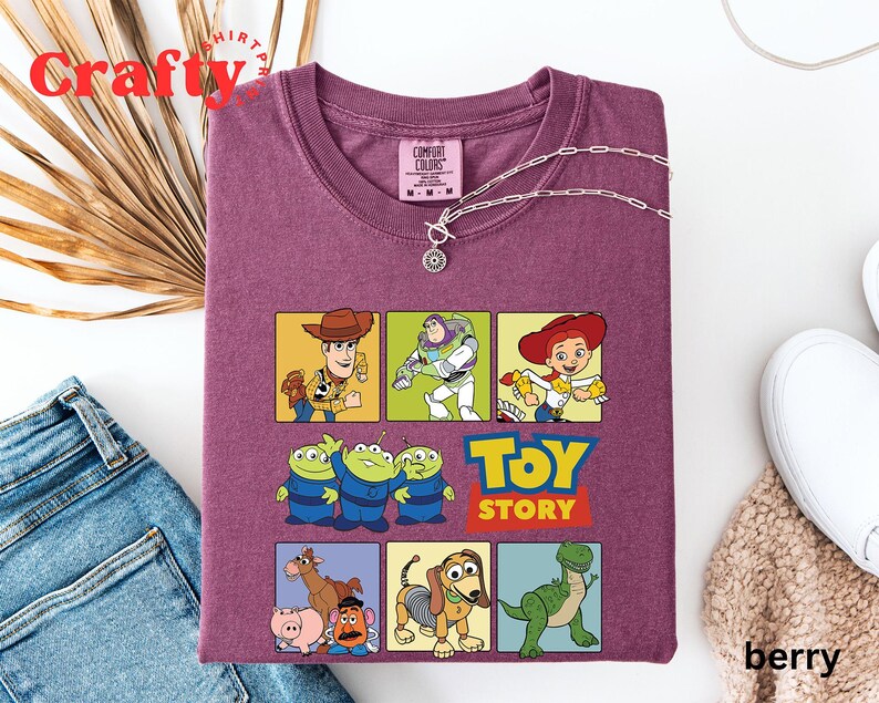 Pu&ograve; includere: Maglietta viola con una grafica di personaggi del film Toy Story. I personaggi includono Woody, Buzz Lightyear, Jessie, i tre piccoli alieni verdi, Rex e Mr. Potato Head. Il testo "Toy Story" &egrave; stampato in lettere bianche su uno sfondo rosso.