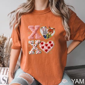 May include: Orange t-shirt featuring embroidered designs. The design includes two "X" letters, one in pink knit and one with black polka dots, and two hearts, one with red hearts and one with a cartoon image of Judy Hopps and Nick Wilde.