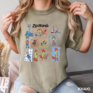 May include: Khaki-colored t-shirt featuring the word "Zootopia" above a grid of cartoon character illustrations. The illustrations include a blue snake, a fox, a bunny, and other characters from the animated movie. The shirt is a comfortable, casual style.