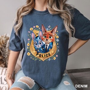 May include: Denim blue t-shirt featuring a colorful graphic of the Zootopia characters Judy Hopps and Nick Wilde inside a police badge. The badge is surrounded by flowers and the word "POLICE". The shirt is a comfortable, casual style.