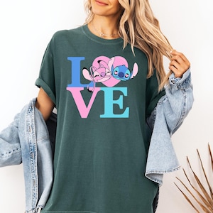 May include: Dark green t-shirt with the word "LOVE" in blue and pink block letters, featuring cartoon characters Angel and Stitch within a pink heart. The shirt is a casual, comfortable style.