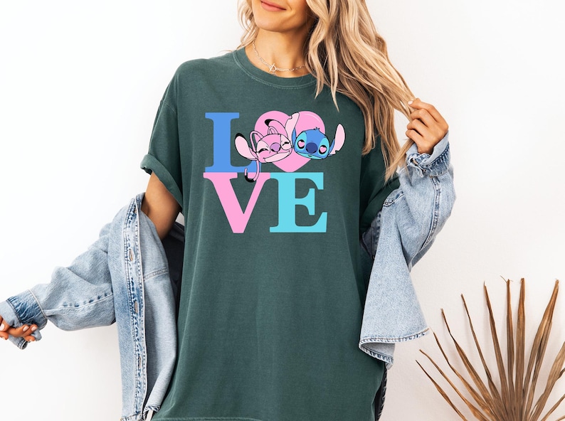 May include: Dark green t-shirt with the word "LOVE" in blue and pink block letters, featuring cartoon characters Angel and Stitch within a pink heart. The shirt is a casual, comfortable style.