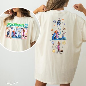 May include: Ivory-colored t-shirt featuring the text "ZOOTOPIA 2" with cartoon characters on the front and back. The back design includes Judy Hopps, Nick Wilde, and other characters from the movie. The shirt is a relaxed fit.