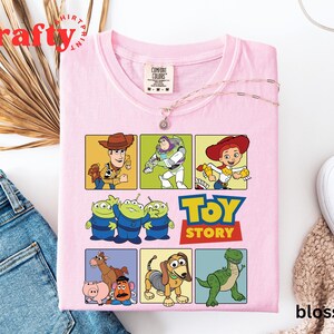 Pu&ograve; includere: Maglietta rosa con tema Toy Story. La maglietta presenta un collage di personaggi del film, tra cui Woody, Buzz Lightyear, Jessie e gli alieni. Il testo "Toy Story" &egrave; stampato in grassetto sulla maglietta.