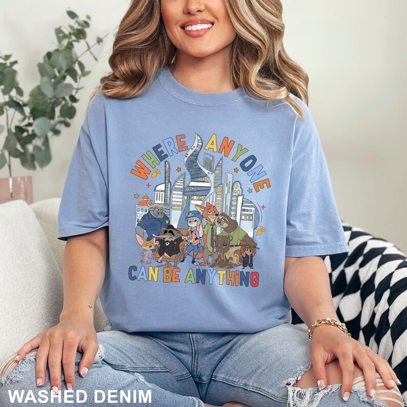 May include: Light blue t-shirt with the text "WHERE ANYONE CAN BE ANYTHING" above a cartoon illustration of various animal characters in front of a city skyline. The shirt is a washed denim color.