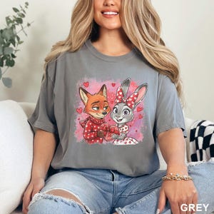 May include: Grey t-shirt with a cartoon fox and bunny design, holding a heart-shaped gift. The fox is in a red scarf and sweater, the bunny in a dress with a bow. Pink background with hearts. The word "GREY" is in the bottom right corner.