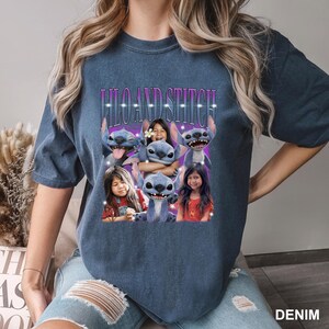 May include: Denim blue t-shirt featuring the text "Lilo and Stitch" above a graphic of the characters Lilo, Stitch, and two girls. The graphic has a purple background with white accents. The shirt is a casual style.