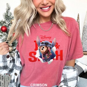 Puede incluir: Camiseta de color carmes&iacute; con un gr&aacute;fico de Disney Stitch. El gr&aacute;fico muestra a Stitch saliendo de un agujero rasgado, sosteniendo un oso de peluche marr&oacute;n. El texto "1 & Stitch" est&aacute; en rojo.