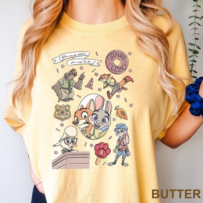 Puede incluir: Camiseta amarilla con personajes de dibujos animados de Zootopia. El dise&ntilde;o incluye a Nick Wilde, Judy Hopps y otros personajes, junto con el texto "You can be whatever you wish to be." La camiseta tambi&eacute;n tiene el texto "THE BIG DONUT."