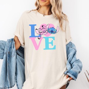 May include: Cream-coloured t-shirt with the word "LOVE" in blue and pink block letters. The "O" is a pink heart with cartoon characters. The shirt is a casual, oversized style.