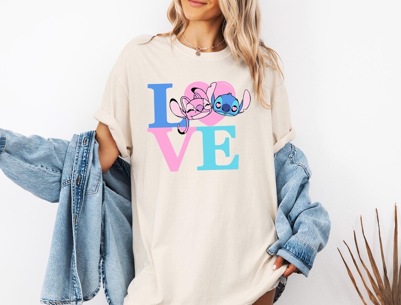 May include: Cream-coloured t-shirt with the word "LOVE" in blue and pink block letters. The "O" is a pink heart with cartoon characters. The shirt is a casual, oversized style.