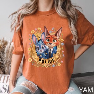 May include: Burnt orange t-shirt featuring a cartoon image of a blue rabbit and a red fox in police uniforms, surrounded by flowers and a gold badge that says "POLICE". The shirt has short sleeves.