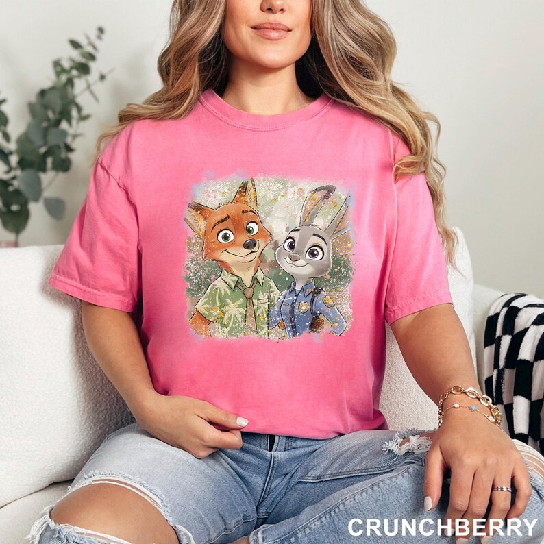 以下が含まれることがあります： ピンク色のTシャツ。映画「ズートピア」のニック・ワイルドとジュディ・ホップスのグラフィックが描かれています。ニックは緑色のシャツを着ており、ジュディは警察の制服を着ています。「CRUNCHBERRY」の文字が画像の下部に印刷されています。